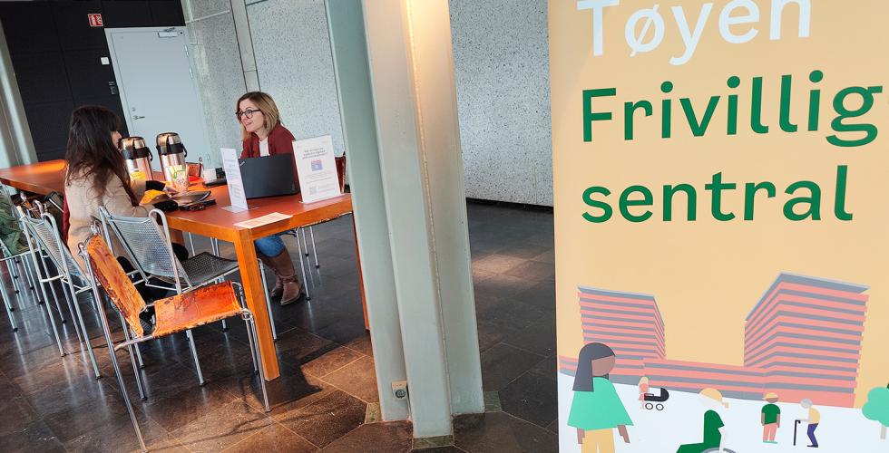 Tøyen Frivillighetssentral som plakat. Sitter to stk ved et oransje bord og prater.