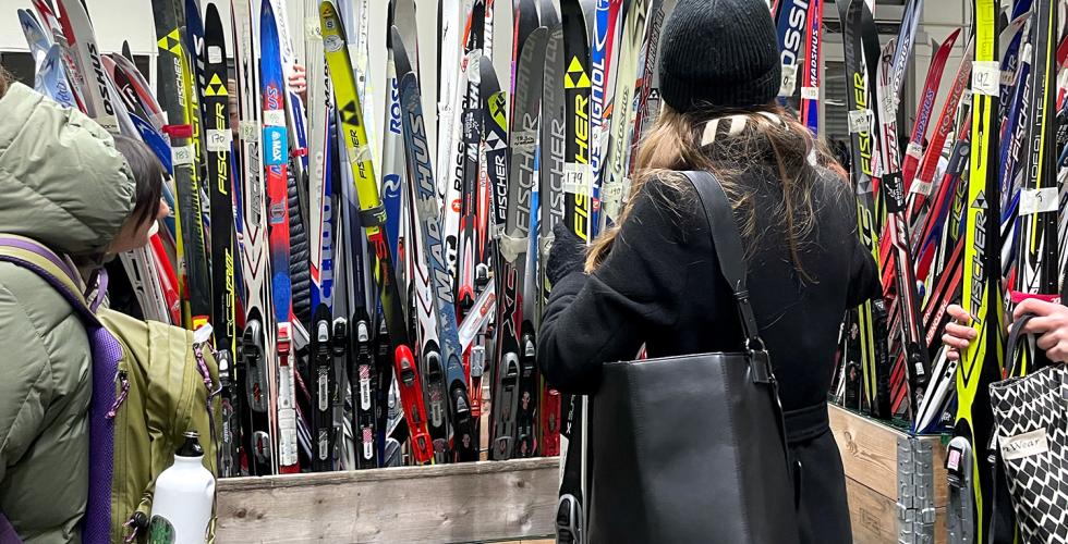 Mange langrennski stå rett opp og ned i en kasse. En kvinne står forran kassen og holder et parr med langrennsski