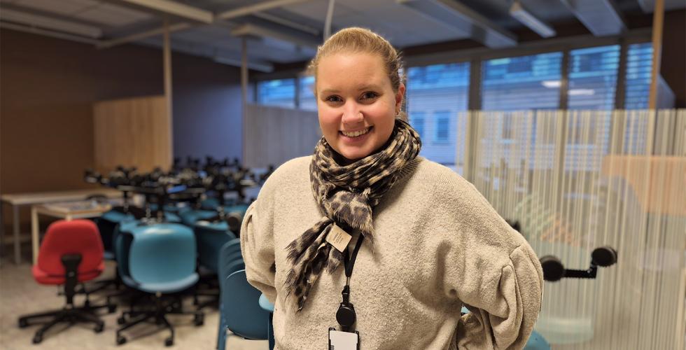 En dame st&aring;r foran et rom under oppussing fylt med stoler. Hun har h&aring;ret opp i en hestehale, og skjerf og genser p&aring;. Hun smiler og har hendene bak ryggen.