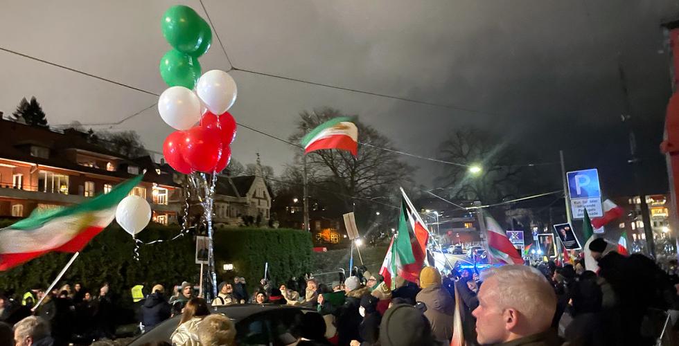 Mennesker st&aring;r langs en gate og holder flagg og ballonger i r&oslash;dt, hvit og gr&oslash;nt. Det er m&oslash;rkt ute