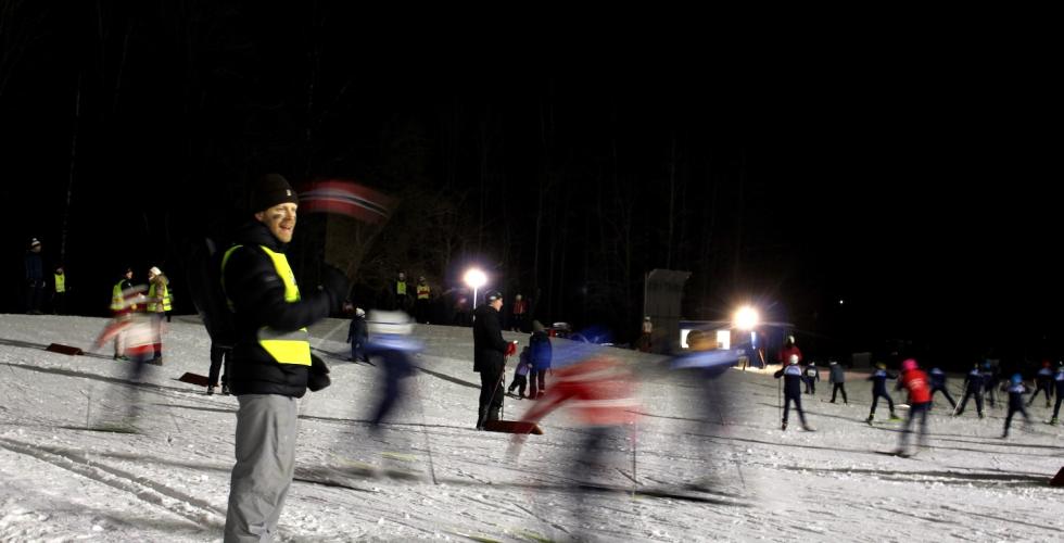 Mann st&aring;r &aring; veiver med norskt flagg foran barn som g&aring;r p&aring; langrenn.