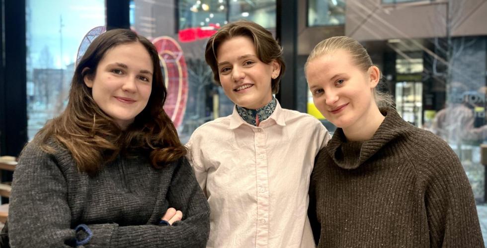 Tre unge kvinner st&aring;r sammen og smiler.