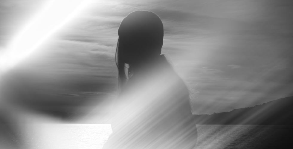 Et sort-hvitt bilde viser silhuetten av en person i profil, st&aring;ende foran et stille vann med lave &aring;ser i bakgrunnen. Mykt, skr&aring;tt lys skj&aelig;rer gjennom bildet og gir en dr&oslash;mmende, nesten t&aring;kete stemning. Bildet er en illustrasjon.