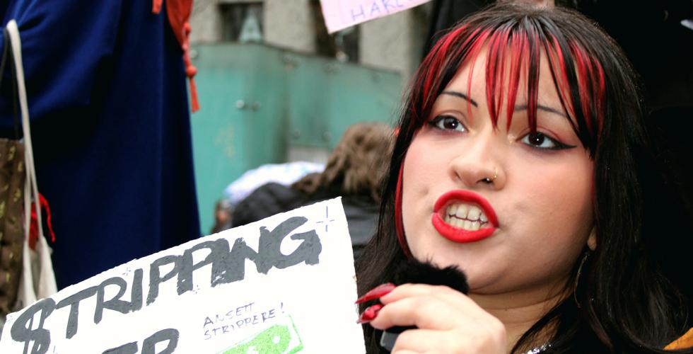 Stripper Alma Adore holder en plakat der det st&aring;r "Stripping er Kunst". Hun er midt i en setning og er derfor hard i fjeset og ser sint ut.