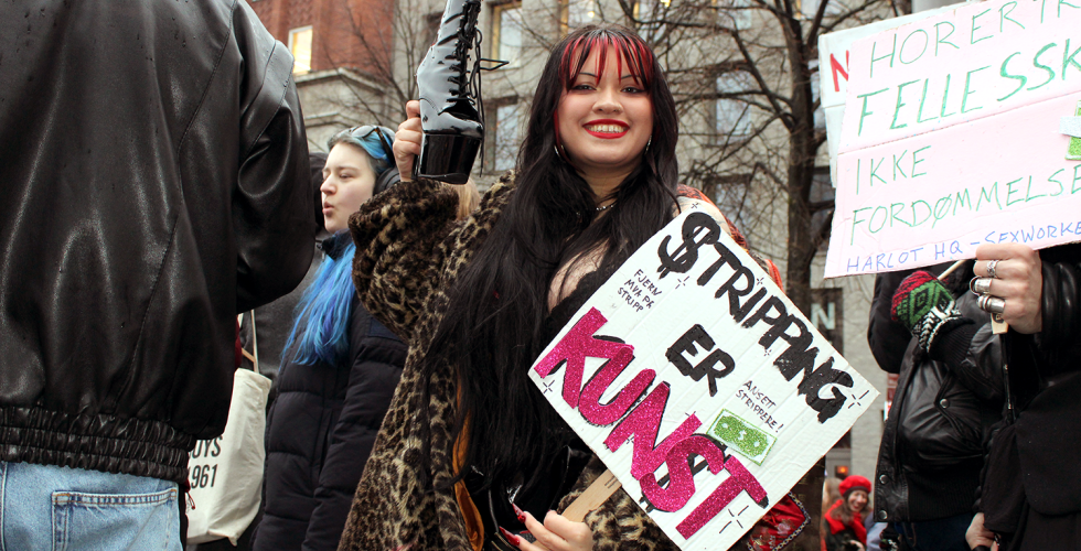 Stripper Alma Adore l&oslash;fter en strippesko i v&aelig;ret med den ene h&aring;nda og holder en plakat der det st&aring;r "Stripping er Kunst" i den andre. Hun smiler til kamera