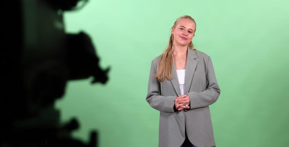 Blond dame st&aring;r forran en gr&oslash;nn vekk et uskarpt kamera er til h&oslash;yre i bildet