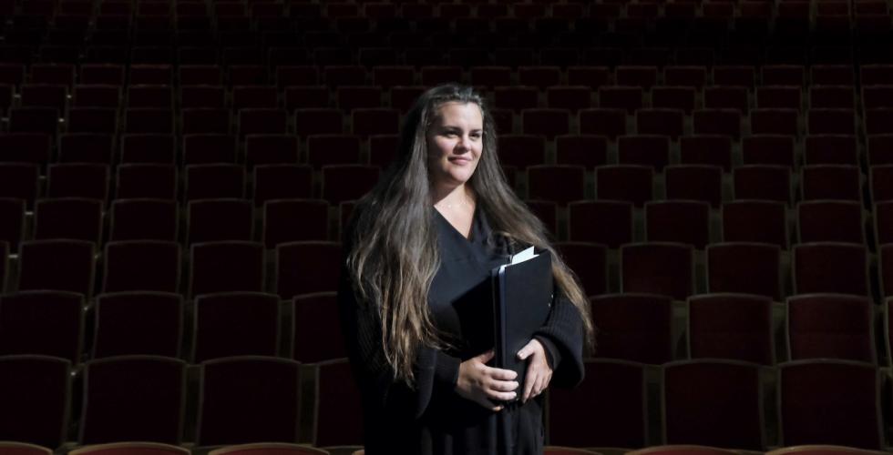Dame står forran en teatersal, scenelyset lyser opp ansiktet hennes