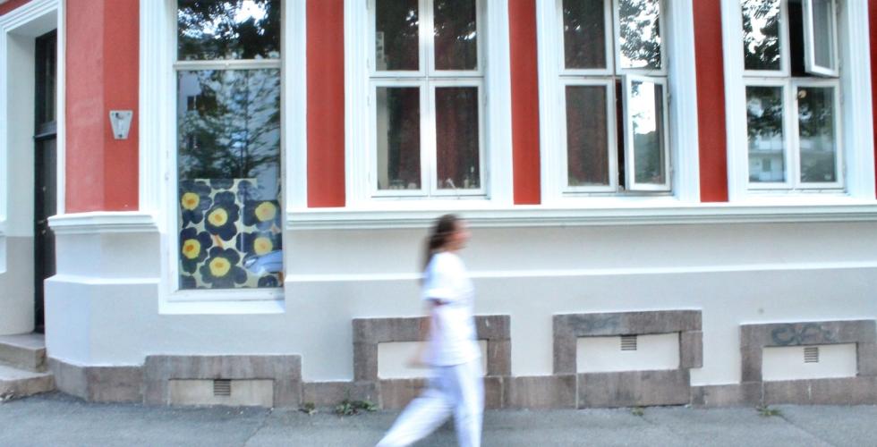 Dame i hvit sykepleieruniform g&aring;r foran en r&oslash;d bygning. Jobber i hjemmesykepleien.