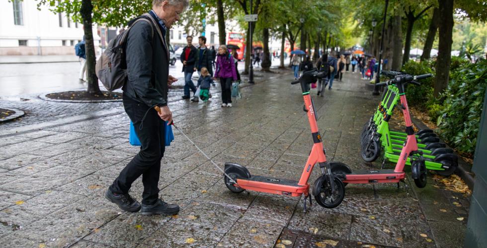 En mann med hvit stokk i høyre hånd går på et bredt fortau. Han møter på en elsparkesykkel som står parkert mer i midten, sammenlignet med resten som står parkert fint til siden. Stokken treffer elsparkesykkelen.