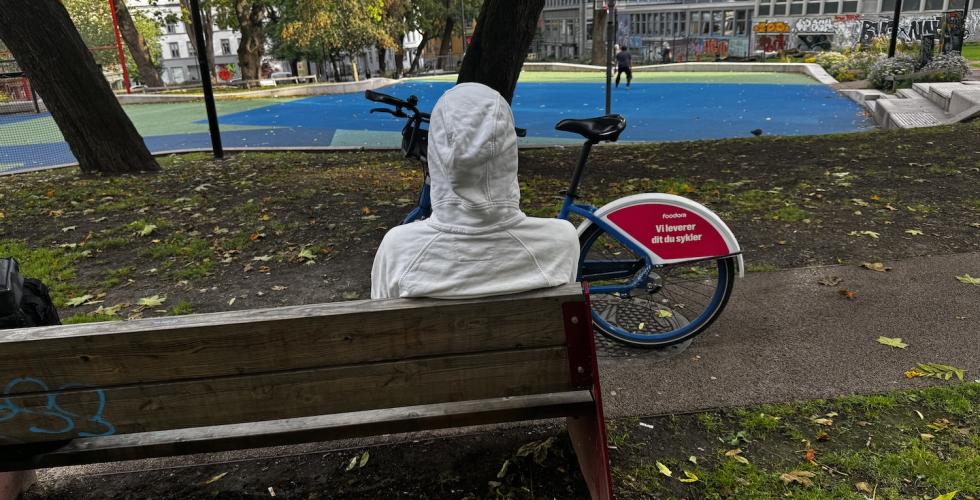 Person med hettegenser sitter på en benk, med ryggen mot fotografen