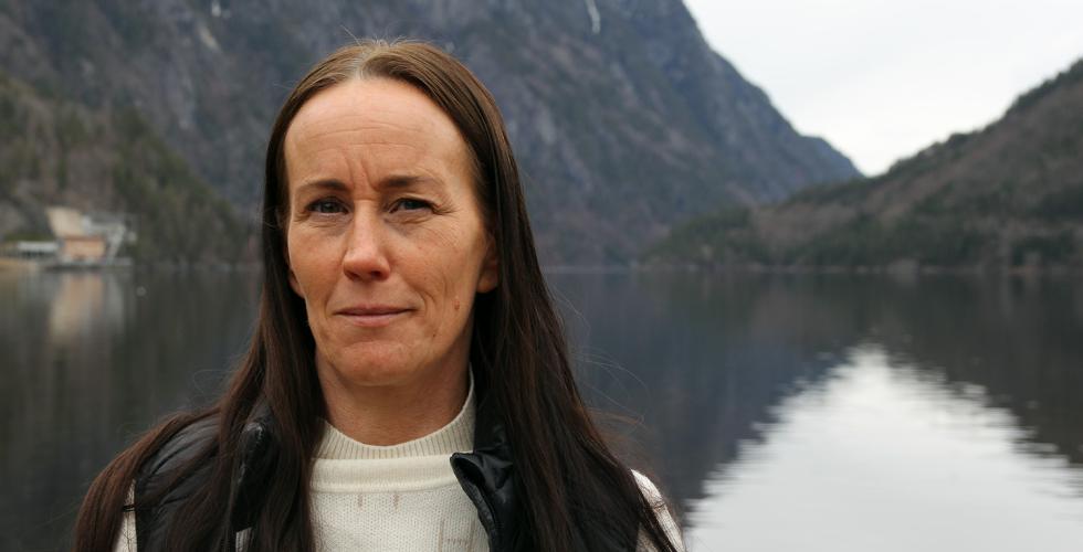 Ann Mari Løvhaug står foran en fjord og to fjell. Ansiktsuttrykket hennes er alvorlig.  