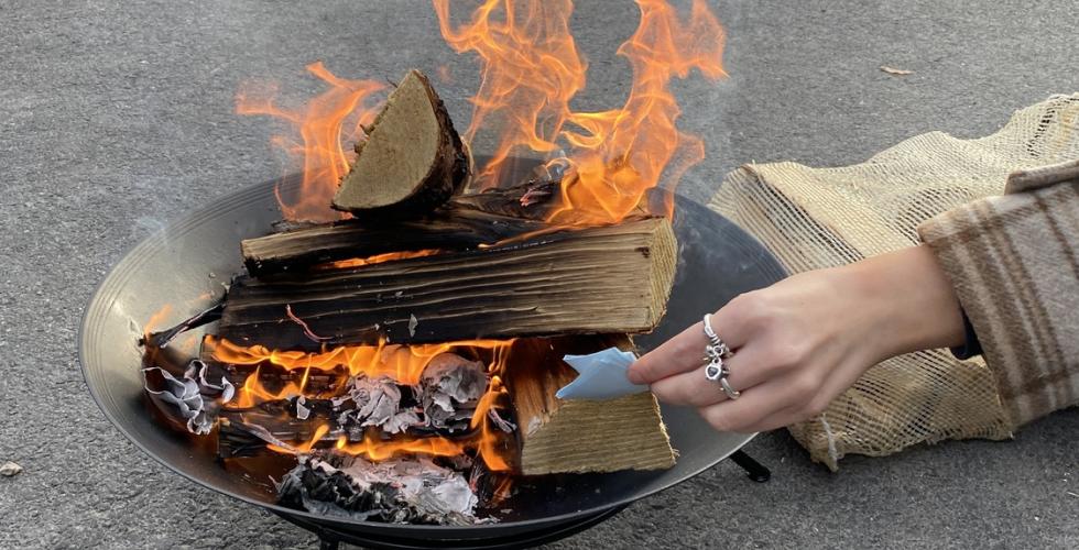 Bilde av et bål som brenner, med en hånd som stikker en lapp inn i bålet.