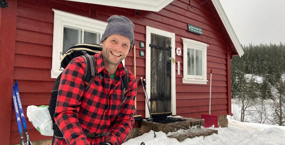 David Brødsgaard smiler og tar på seg skiutstyr foran den røde hytten Skjennungsvolden. Det er snø rundt ham. 
