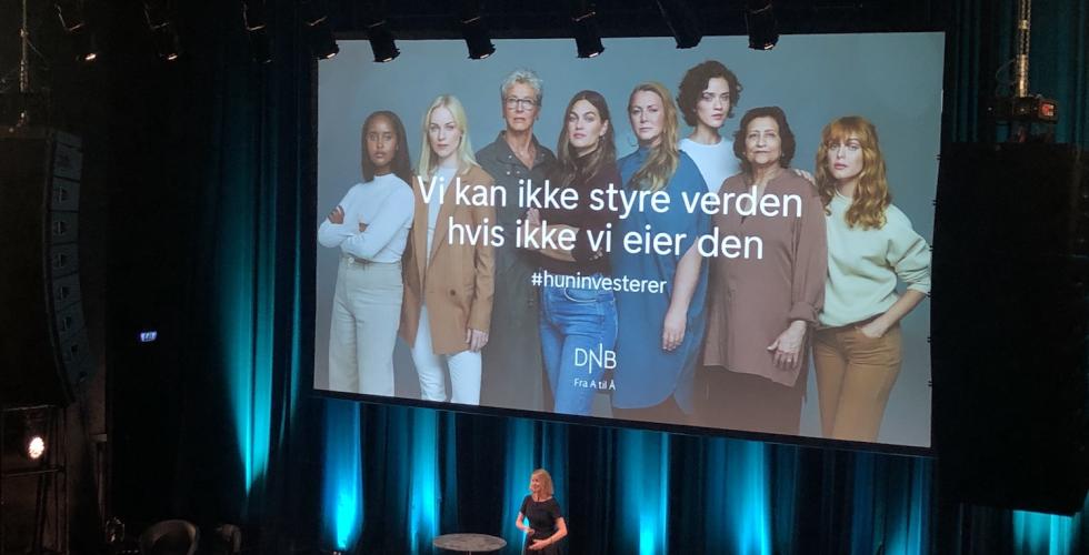 Aina Lemoen Lunde foran et stor publikum på Sentrum Scene i Oslo, med DNBs reklame for #HunInvesterer på lerret bak seg.