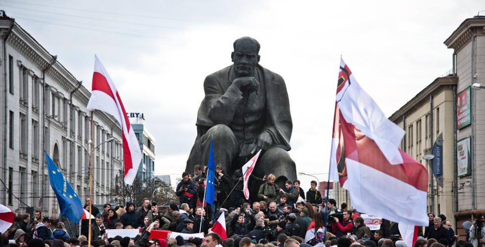 Mange demonstranter som bærer på hviterusslands hvite og røde flagg. De er vendt med ryggen mot kameraet og ser på en stålstatue av Lenin.