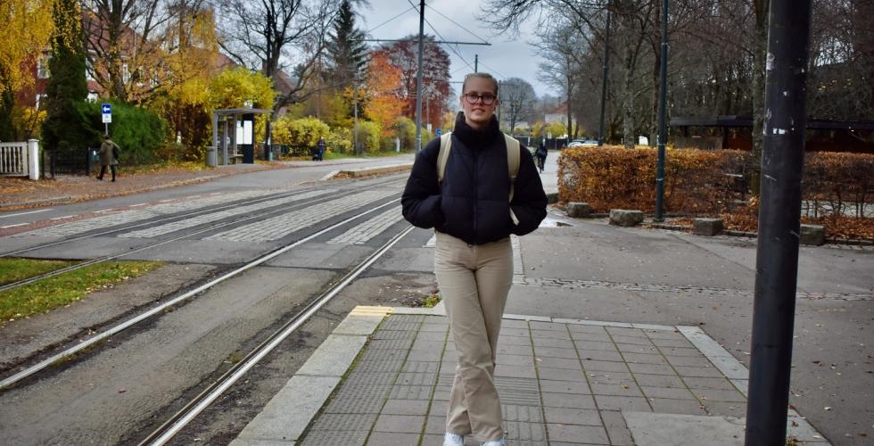 Blond kvinne med svart jakke, beige ryggsekk og mørke briller står og venter på trikken utenfor Universitetet i Oslo, avd. Blindern