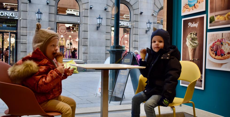 To små barn i parkdresser og luer sitter inne på en iskrembutikk og spiser is en kald novemberdag. Begge spiser en kule sjokoladeis i kjeks. Gjennom vinduet utenfor kan man se travle mennesker på Karl Johan. 