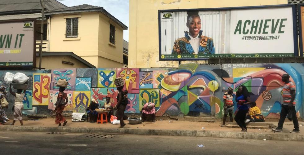 Bildet viser et fortau i Ghana. Det går flere personer på fortauet, og bak dem er en vegg dekket i fargerik graffiti. 