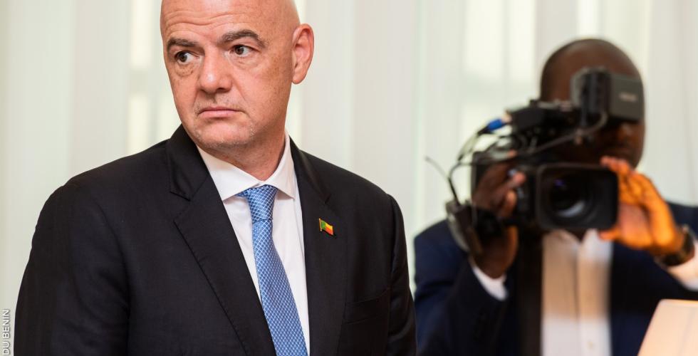 Bilde av Gianni Infantino i en svart dress med blått slips og en kameramann i bakgrunn.