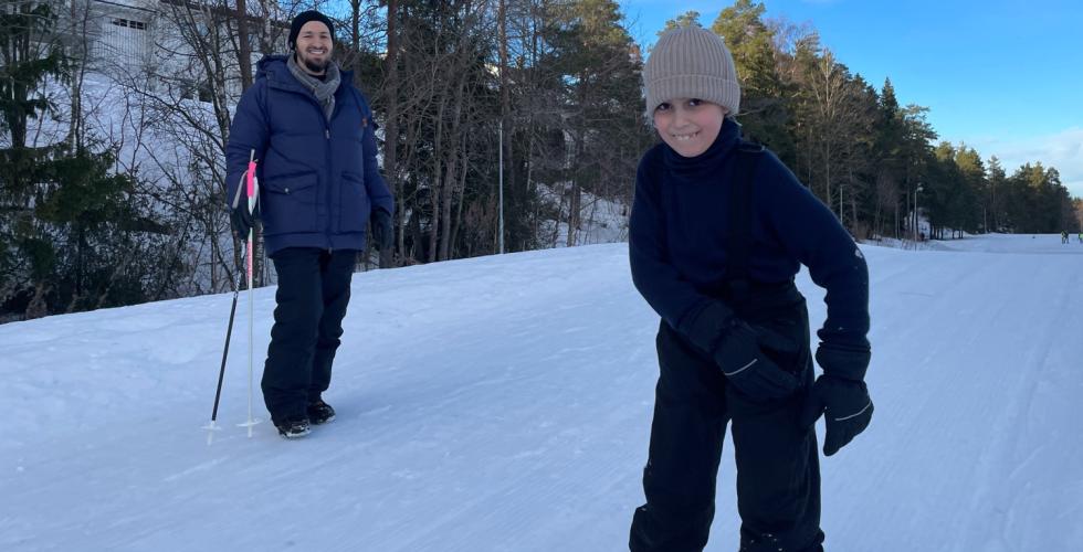 Gutt som står på ski med faren i bakgrunn.