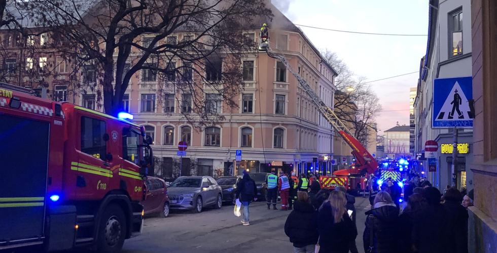 Bygningen det brant i i Møllergata, tatt fra fortauet over veien litt bortenfor bygget. Mørkegrå røyk stiger ut fra et vindu i 4. etasje, der vinduene er borte og det ser helt svart ut inni. Bortsett fra det ser bygårdens fasade ganske dagligdags ut.