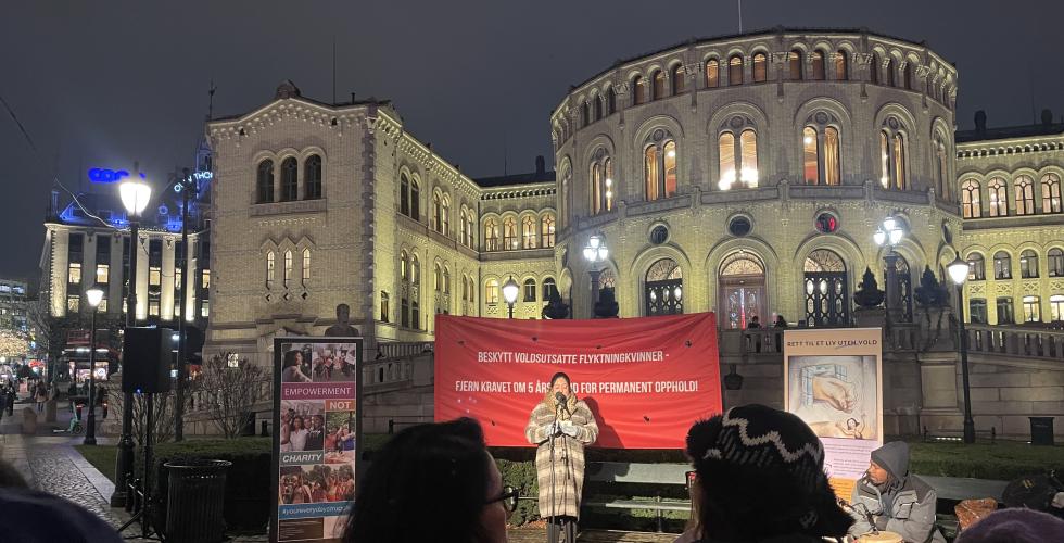 Bildet viser Khansa Ali som står foran Stortinget og holder en appell. Bak henne henger det et rødt banner hvor det står "Beskytt voldsutsatte flyktningkvinner - fjern kravet om 5 års botid for permanent opphold".