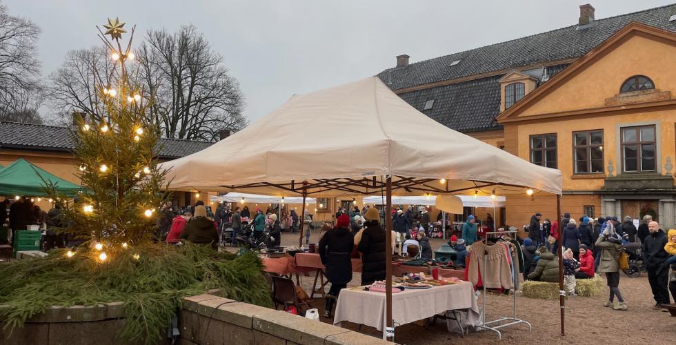 Bildet viser julemarkedet på Bogstad Gård. Man kan se et juletre med lys, et hvitt telt som holder regnet unna noen som selger produkter, og mange folk i bakgrunnen. 