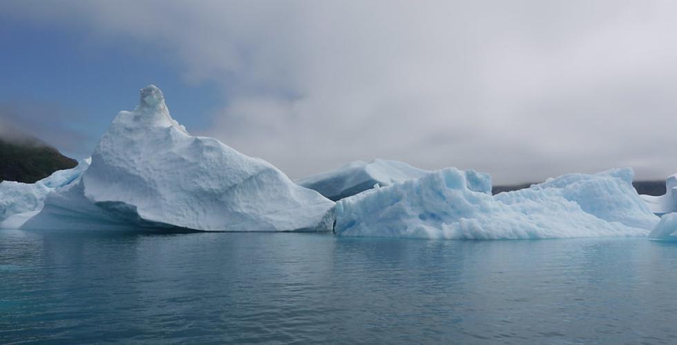 Bildet viser en liten isbre på Grønland, liggende på et stille turkist hav, over isbreen er det mørke skyer