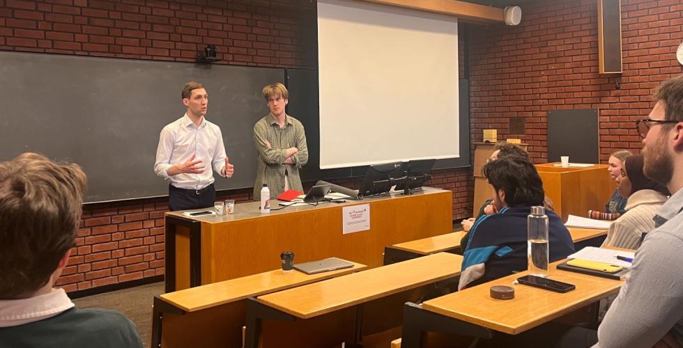 En mann i hvit skjorte står foran i et auditorium sammen men en gutt i grønn skjorte. Studenter følger med fra salen. 