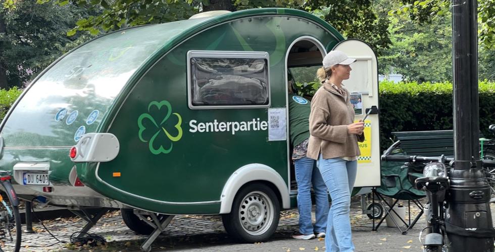 Bilde av en blond kvinne i caps, beige jakke og lyseblå jeans foran senterpartiet sin grønne campingvogn-valgbod på Karl Johan, som ser til siden