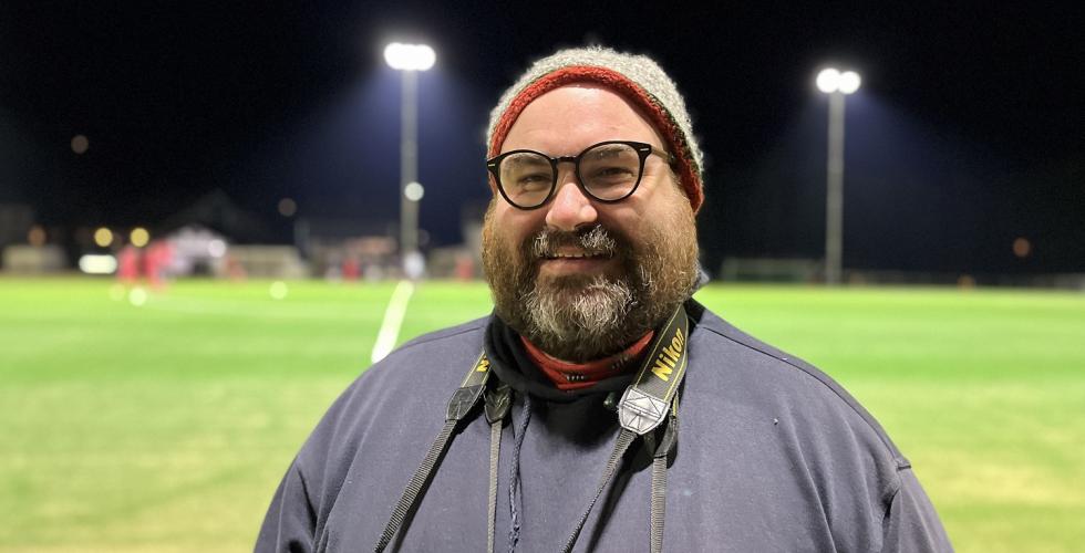 Marius Helgå smiler til kamera, med en fotballbane i bakgrunnen.