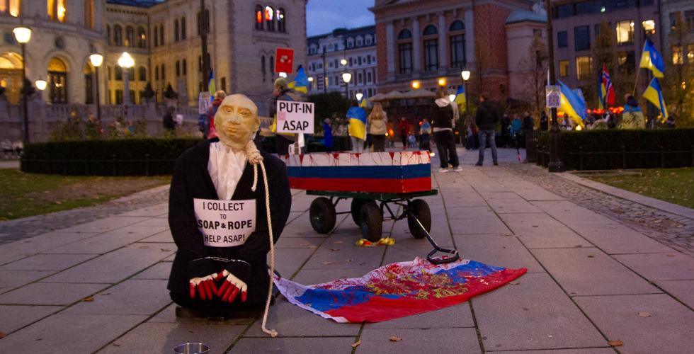 En dukke som forestiller Putin står på kne foran en metallskål. I bakgrunnen står en kiste på hjul malt etter det russiske flagget. 