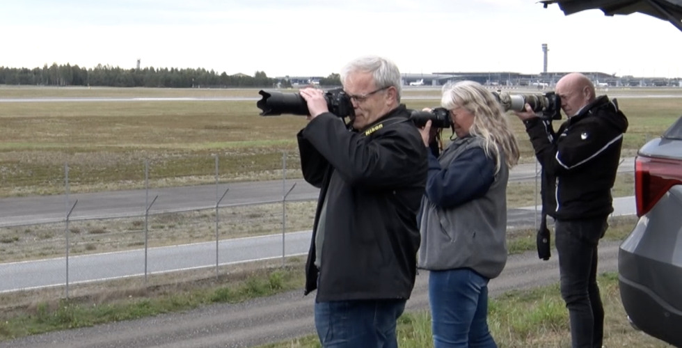 Tre flyspottere med kameraet mot ansiktet i høyre hjørnet av bildet. Rullebane på Gardermoen i bakgrunnen