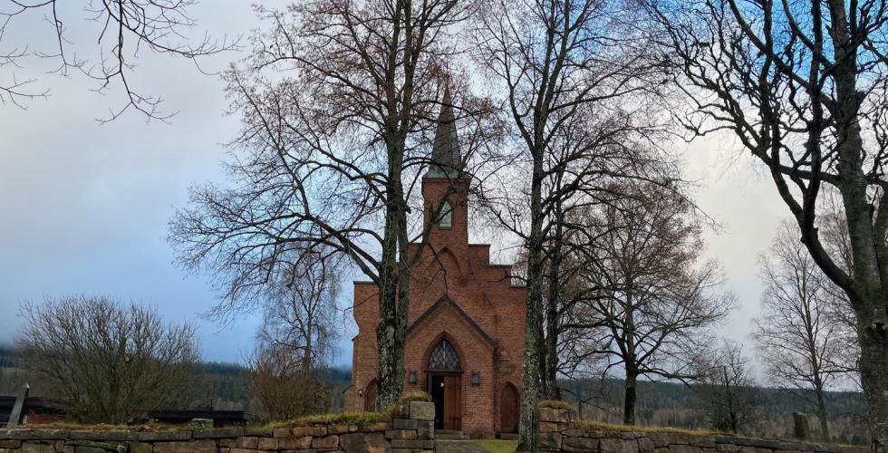 Bildet viser Sørkedalen kirke i Oslo. Kirken er laget av brune murstein, og har et høyt kirkespir. Den står litt tildekket av nakne løvtrær. 