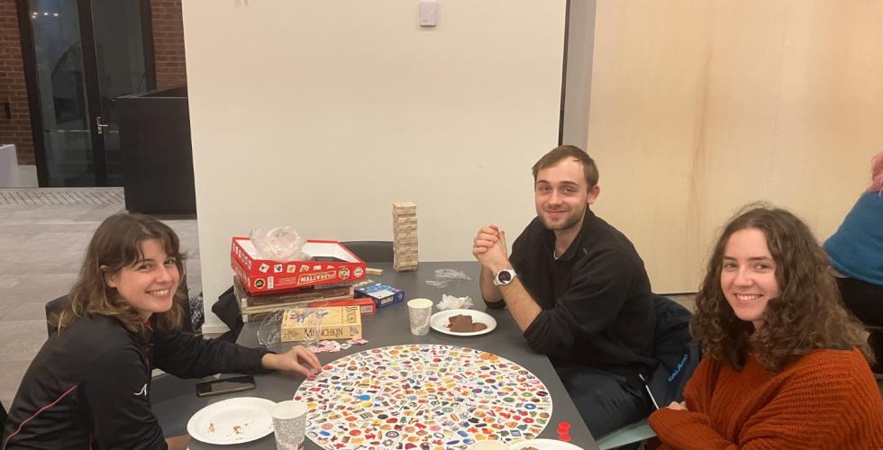 Bord med tre studenter rundt, på bordet ligger noen brettspill og papptallerkner med snacks