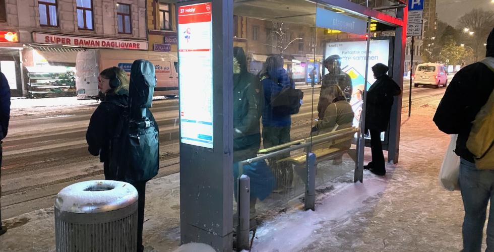 Bildet viser flere mennesker som venter på bussen på Tøyengata buss-stopp, vendt mot Super Bawan Dagligvare. Det er litt mørkt ute, og snøer faller ned. Det er snø på bakken. Personene står med ryggen til fotograf.