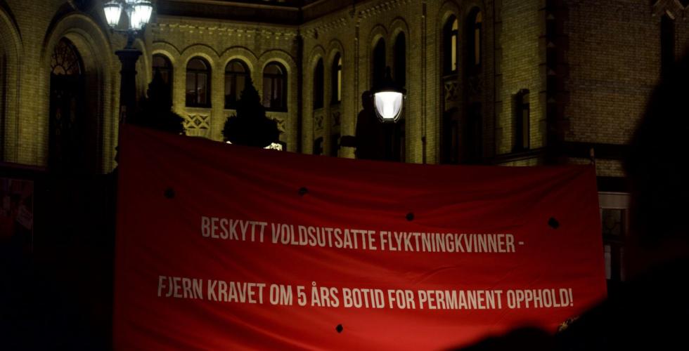 Rød banner på plassen foran Stortinget der det står beskytt voldsutsatte flyktningkvinner fjern kravet om fem års botid for permanent opphold