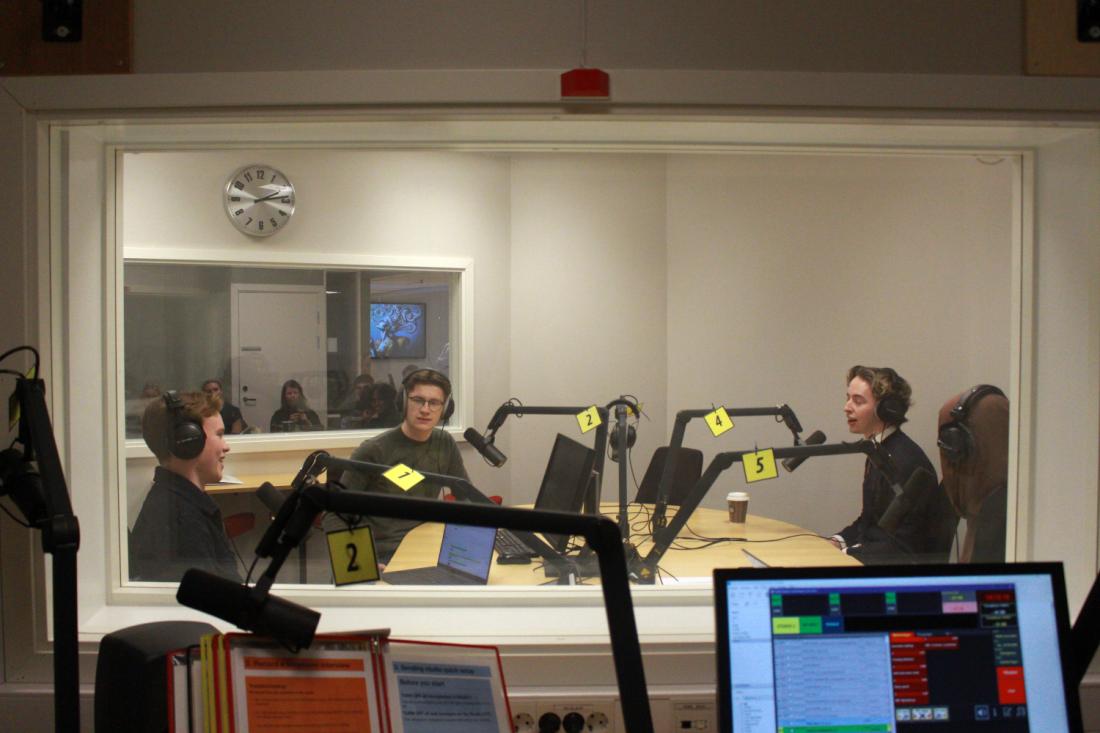 Fire mennesker inni et radiostudio gjennom et vindu 