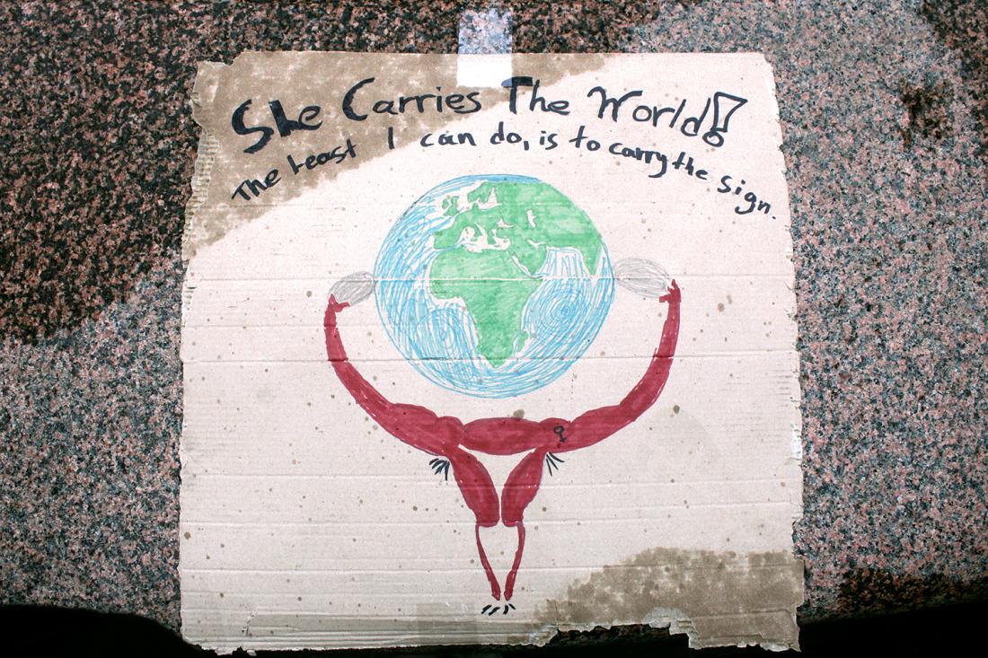 Plakat der det st&aring;r She carries the world - the best I can do is to carry the sign, med bilde av en livmor som b&aelig;rer verden